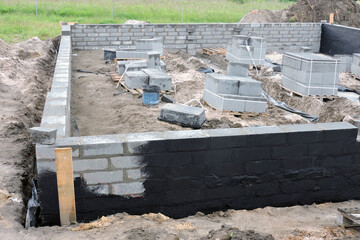 Damp proofing the outside foundation wall with a black black asphalt-based mixture, a construction site with solid concrete blocks on pallets, plastic buckets and mixing tubs