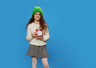 Portrait of a cheerful little girl with a green hat and wavy hair, modern trend, isolated on a blue background, holing a red alarm clock.
