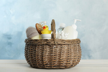 Wicker basket full of different baby cosmetic products, accessories and toy on white wooden table