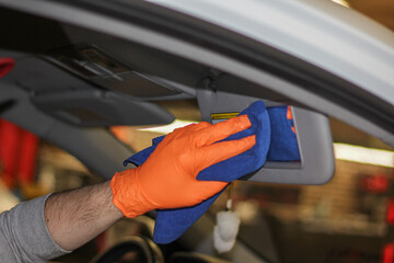 Detailing and car cleaning services, the concept of car washing and cleaning. A male worker in orange rubber gloves is cleaning the car sun visor mirror with a blue microfiber towel.
