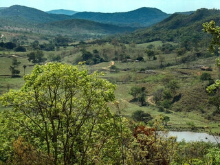 Linda paisagem na região rural de Esmeraldas, onde vemos ao longe, muita vegetação, um pequeno lago, construções, gado no pasto e uma estrada de terra que corta uma fazenda.