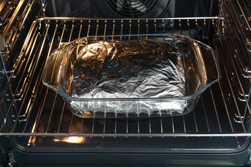 Glass tray with piece of meat wrapped in aluminum foil baked  in the oven. - obrazy, fototapety, plakaty