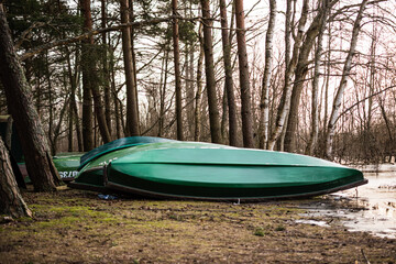 Several green upturned fishing boats lie on the shore near a lake in a mixed forest in Latvia.