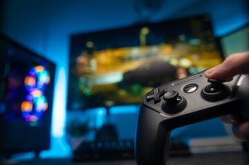 Modern equipment for video games. Gamepad in the gamer's hand, large TV screen, personal computer monitor. Blue neon lighting. Video games, entertainment, youth culture.