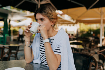 European woman with light hair wearing striped t-shirt and blue shirt is drinking morning coffee in outdoor cafe in morning 