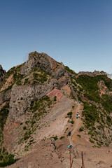 High wild mountains are covered green bushes against blue sky. Path among mountains. People are going on the road. People conquer the mountains