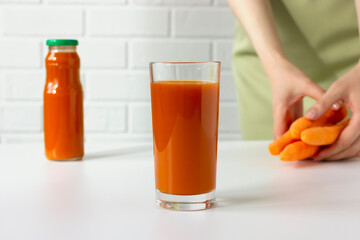 Glass of fresh carrot juice, bottle of carrot juice, raw carrots holding by a woman on the kitchen, horizontal plane