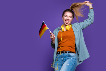 Girl in casual clothes makes funny faces, shows tongue and holds small German flag. Study abroad...