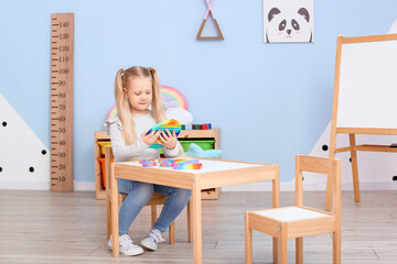 Pretty little girl sitting at table and playing with pop it fidget toy in child room - obrazy, fototapety, plakaty