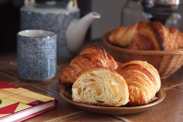 A cooked traditional French butter croissants with its cut piece, it's a fluffy of multiple layers. With teapot and cup in background. It's a buttery, flaky pastry fold with butter in multiple layers 