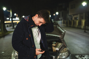 Fototapeta na wymiar One young adult caucasian man standing by his vehicle with open hood and broken failed engine holding a phone calling towing service for help on the road Roadside assistance concept in dark night