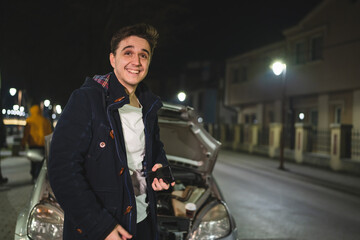 One happy young adult caucasian man standing by his vehicle with open hood and broken failed engine smiling carefree hold phone calling towing service for help on the road Roadside assistance concept