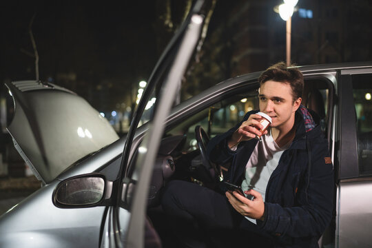 One Young Adult Caucasian Man Sitting His Vehicle With Open Door And Hood And Broken Failed Engine Holding A Phone Calling Towing Service For Help On The Road Roadside Assistance Concept In Dark Night