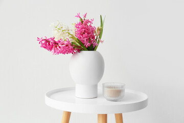 Vase with hyacinth flowers and candle on table near white wall