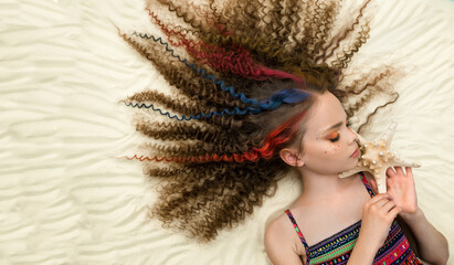 Cute little child girl with long curly colored hair lays on the sands background. Face of beautiful teen girl with curly hairstyle posing with seashells on the sand. Top view