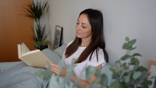  A beautiful woman reading a book and relaxing on the bed in the bedroom in the morning. Resting on the weekend with a book in a calm environment.