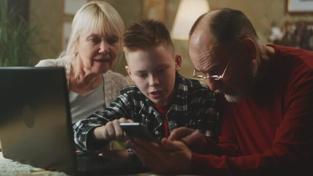 Grandmother laughing and watching teenager teaching grandfather using cellphone and netbook in living room at home