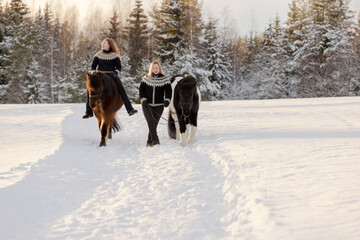 Fototapeta na wymiar Two Icelandic horses and riders walking on the snowy road in countryside. During sunset.