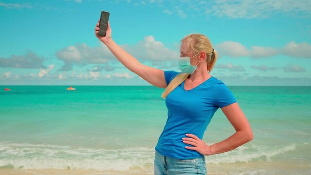 blonde girl in a mask coronovirus look at the phone on the shore blue sea