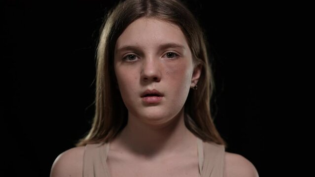 Close-up front view portrait of Caucasian teenage girl opening face showing bruises looking at camera. Sad abused teenager posing at black background with desperate facial expression