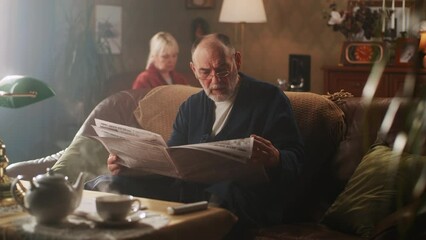 Pan left view of aged man in bathrobe and glasses sitting on couch and reading daily newspaper in cozy living room, near woman in morning at home - Powered by Adobe