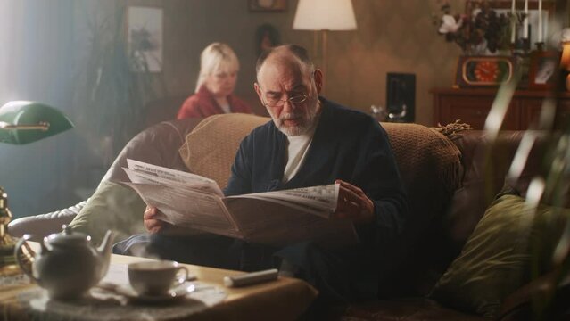 Pan left view of aged man in bathrobe and glasses sitting on couch and reading daily newspaper in cozy living room, near woman in morning at home