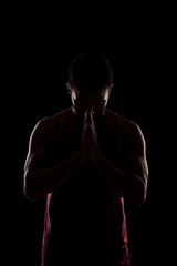 Fototapeta na wymiar Side lit muscular Caucasian man silhouette. Athlete in red shirt praying against black background