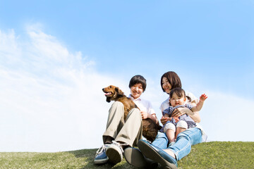 青空を背景にペットの犬を抱き芝に座る笑顔の若い夫婦と幼い娘。家族,幸せ,愛情,育児とペットのイメージ