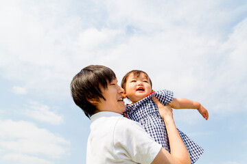 青空を背景に幼い娘を抱き上げる若いお父さんをローアングルで見上げる。親子,家族,父子,育児のイメージ