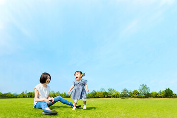 青空を背景に緑の芝生の上で楽しく遊ぶ母と幼い女の子。親子,幸せ,愛情,育児のイメージ