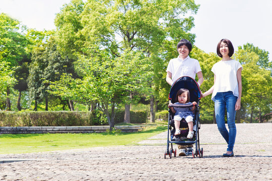 青空を背景に緑豊かな公園の遊歩道をベビーカーを押しながら散歩をする若い夫婦。幸せ,家族,愛情,育児のイメージ