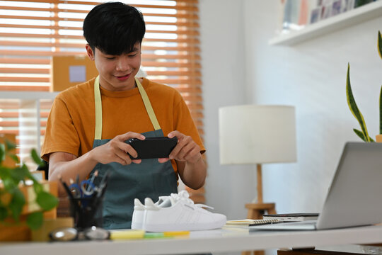 A portrait of a young handsome Asian e-commerce employee standing in the office full of packages in the background taking a picture of a product, for SME business, e-commerce and delivery business.