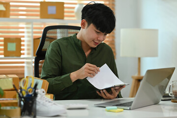 A portrait of a handsome Asian man, e-commerce employee sitting in the office full of packages in the background holding papers andusing a notebook, for SME business, e-commerce and delivery business.