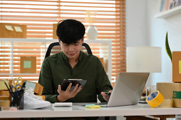 A portrait of an Asian man, e-commerce employee sitting in the office full of packages in the background checking on the document and smiling, for SME business, e-commerce and delivery business.