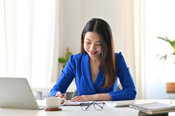 A portrait of a young pretty Asian woman with a suit sitting in the office using a laptop, calculator, working on data and documents on the table, for business, finance and technology concept.