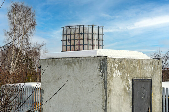 A snow-covered outbuilding with an empty water tank on the roof on a winter day