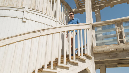 Successful man in blue tennis shirt on vacation descends white spiral stairs at beach luxury...