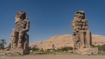 Giant stone sculptures of the Colossi of Memnon in Egypt. Huge statues of seated pharaohs against a clear blue sky and sand dunes.
