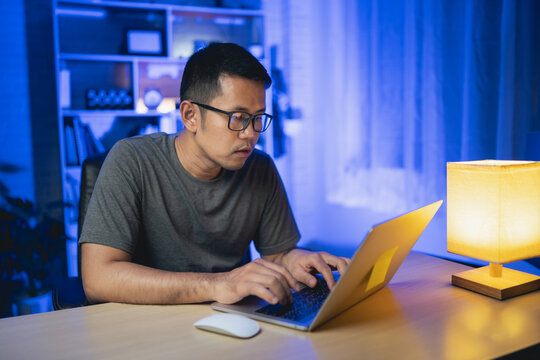 Freelance Man Working On Laptop At Home At Night. Man Freelancing, Thinking, Serious While Working At Home At Night. Freelance Work And Work From Home Concept.