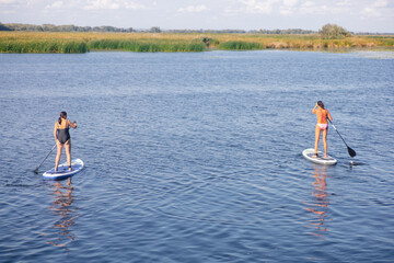 Two middle aged ladies on sup boards each holding oar in hands somewhere on blue lake with reeds and trees far away in background wearing swimming suits. Active lifestyle for older people. 