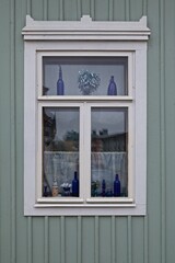 White framed window on a green wooden house. The window has lace upholstery and embellishments.