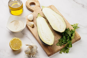 Ingredients for a traditional dish of arabic cuisine baba ganoush: cut in half eggplant, tahin, cilantro, garlic, olive oil, lemon juice on wooden board