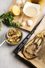Ingredients for a traditional dish of arabic cuisine baba ganoush: roasted eggplant in bowl, tahin, cilantro, garlic, olive oil, lemon juice on wooden board and baking tray