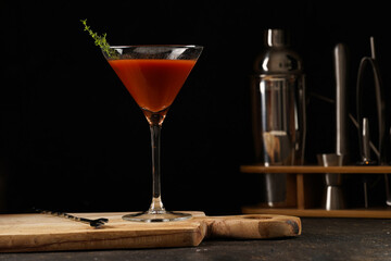 Red non-alcoholic tomato cocktail in martini glass on wooden board on black background and bar equipment