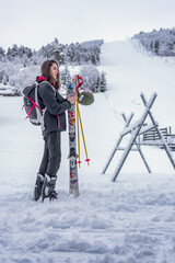 Woman with brown hair, ski wear and backpack preparing for skiing, standing in the valley, of a snow landscape, holding her ski equipment, vertical