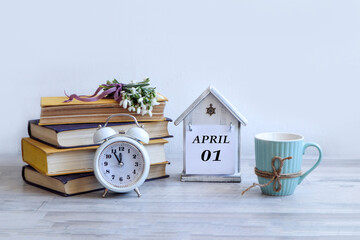 Calendar for April 1: a decorative house with the name of the month April in English, the numbers 01, a stack of books, a bouquet of snowdrops on them, an alarm clock