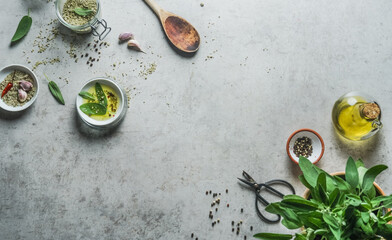 Food background with  flavorful ingredients: herbal salt, sage, olive oil, spices, wooden cooking spoon and kitchen scissors at grey concrete table. Top view.