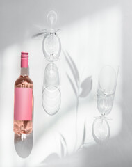 Rose wine bottle with empty pink mock up label and two wine glasses at white background with sunlight and shadow. Modern wine concept. Top view with copy space.