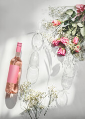 Obraz na płótnie Canvas Rose wine bottle with empty pink mock up label, two wine glasses and romantic flower bouquet on white background with sunlight and shadow. Modern wine concept. Top view with copy space.