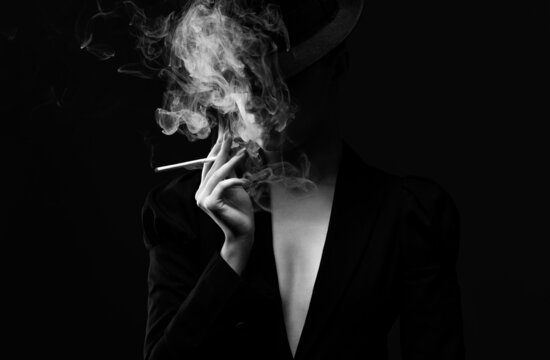 A mysterious woman in a black suit and hat smokes a cigarette. Black and white photo.